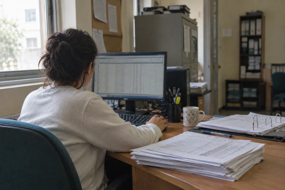 Una persona de espaldas frente a la computadora, con una pila alta de formularios impresos ocupando el escritorio.